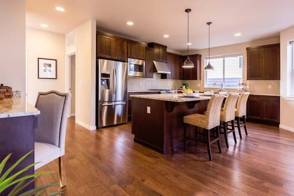 A modern kitchen with a center island that has a sink on it. The kitchen is well lit by the recessed lighting in the ceiling. There are four chairs around the table which have beige cushions on them. Above the stove there is a hood and above the oven there is a range hood.