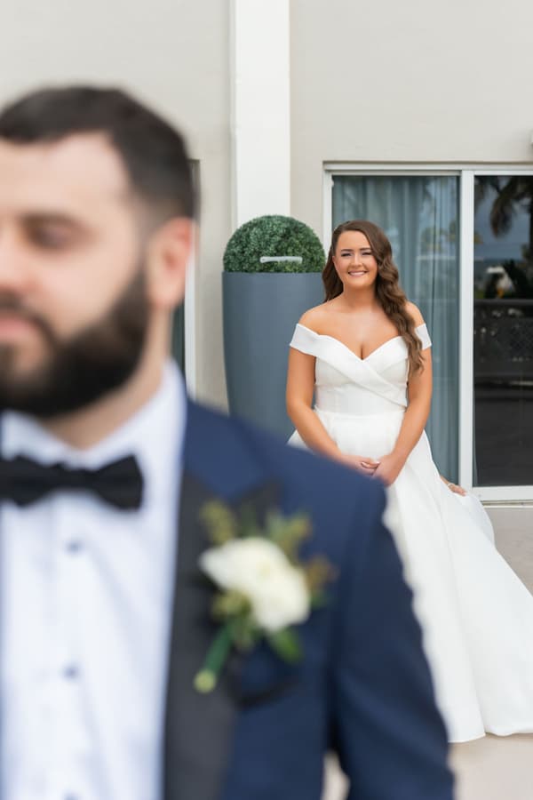 A bride in a white off-the-shoulder gown smiles at her groom dressed in a classic tuxedo during their wedding first look.