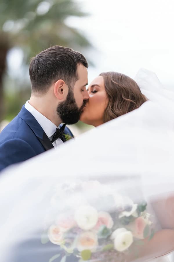 A tender moment of a bride and groom sharing a kiss under the veil, set against an overcast sky with soft natural light.