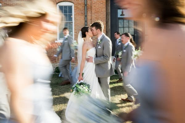 A bride and groom share a kiss amidst blurred figures in an outdoor wedding ceremony.