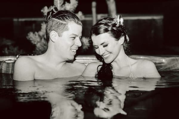 A man and a woman are sitting in a pool together with smiles on their faces. The photo is black and white.