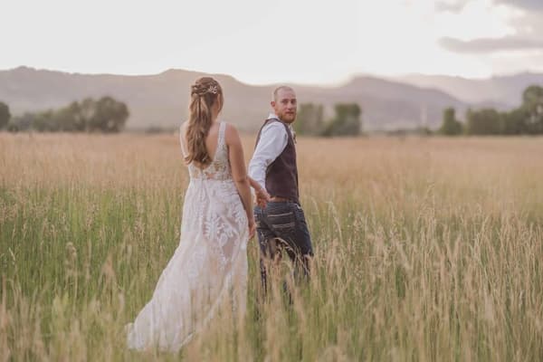 A man and a woman are standing in the middle of a field with tall grass. The woman is wearing a white dress and has her back turned to the camera while the man is looking at her. They seem to be enjoying their time together in this serene setting.