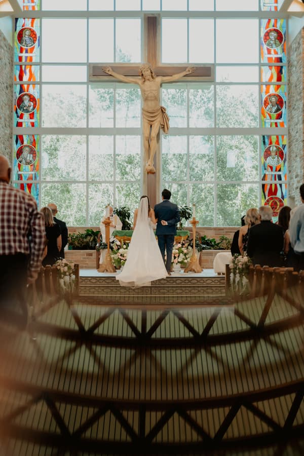 A man and woman stand before a cross with their wedding party behind them.