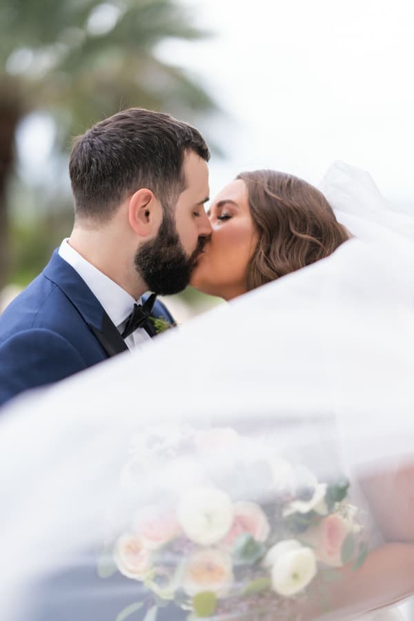 A tender moment of a bride and groom sharing a kiss under the veil, set against an overcast sky with soft natural light.