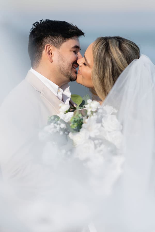 A bride and groom share a tender kiss against a soft, ethereal backdrop of misty clouds during golden hour.