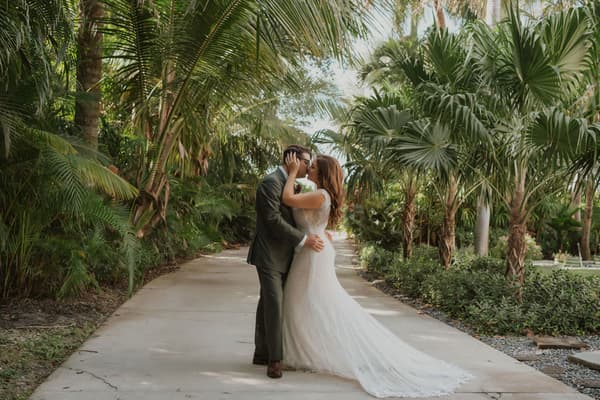 A bride and groom share a tender kiss under the lush canopy of palm trees in an outdoor garden setting.