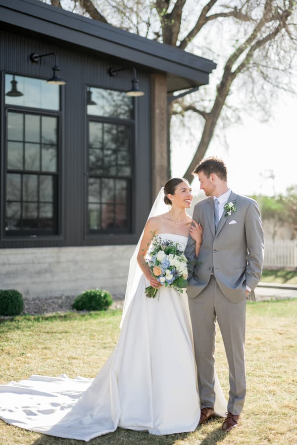 * A bride and groom standing together outside of a building. The man is wearing a gray suit with white flowers on his lapel. The woman is wearing a long white dress with greenery in her hair. * They are holding hands and smiling at each other. * Behind them, there is a black building with windows and lights hanging from the ceiling