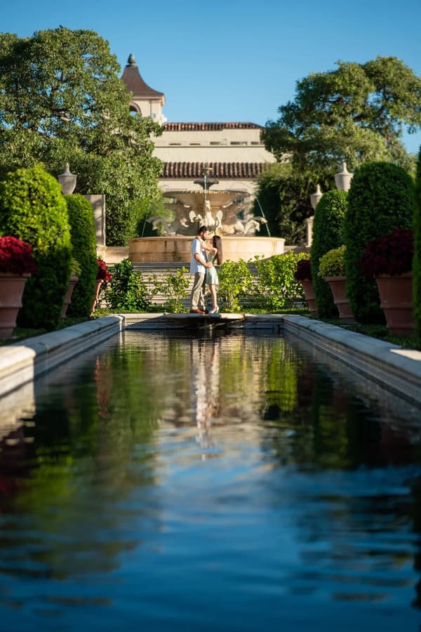 A couple stands embraced on a stone bridge amidst lush greenery and a grand fountain in an elegant outdoor garden setting.