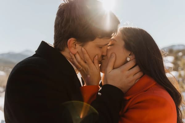 A tender moment of two individuals sharing a kiss under the golden glow of sunlight with a scenic mountain backdrop.