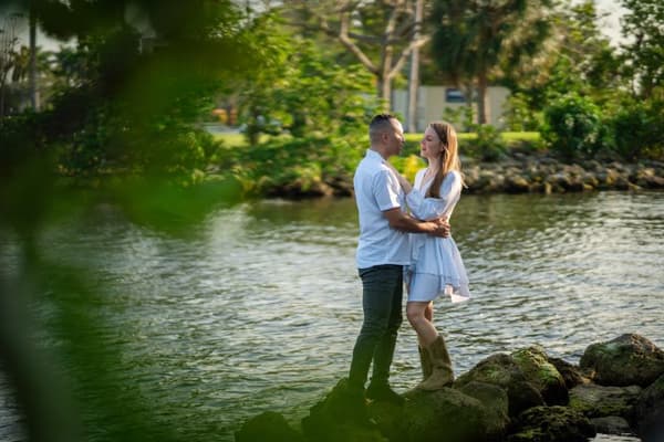 A couple standing on rocks by a body of water with trees behind them.