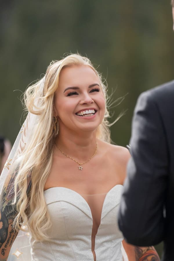 A blonde woman wearing a white dress with tattoos on her arm is smiling at the camera.