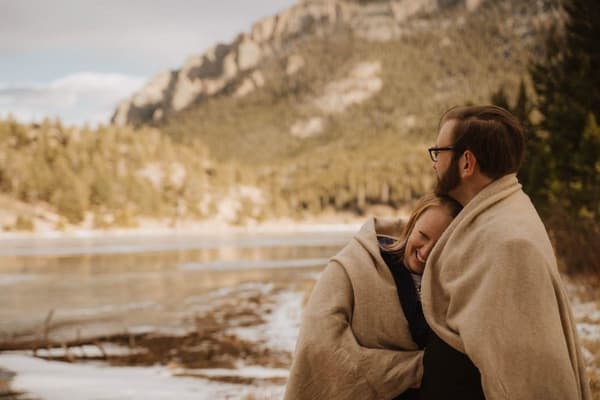 A man with glasses is holding a woman wearing a brown shawl on his shoulders while they look at the water together.